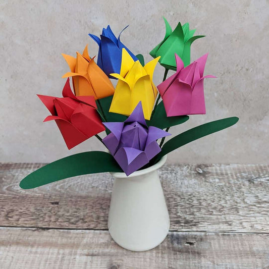 A bouquet of seven origami paper tulips in rainbow colours in a cream vase