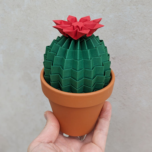 large dark green origami cactus plant with a red paper flower on top, presented by a hand holding the terracotta pot