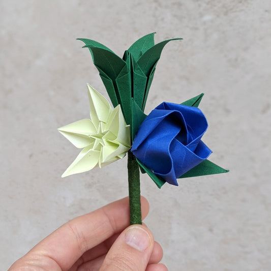 origami buttonhole with three paper flowers: a dark green lily, a blue rose and a pale green star flower