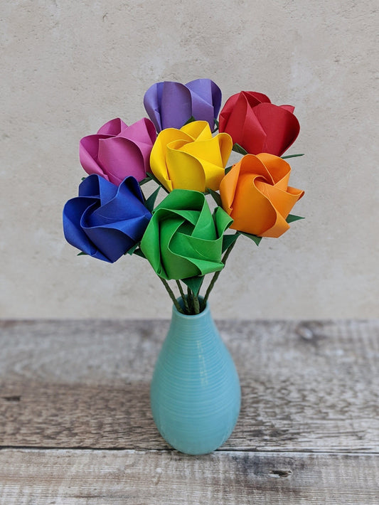 A bouquet of seven origami roses on long green stems in a pale blue vase. The roses are folded from recycled paper and there is one of each colour of the rainbow: red, orange, yellow, green, blue, pink and purple