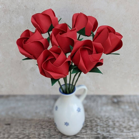 Seven red origami paper roses gathered together into a bouquet. They stand in a small white vase