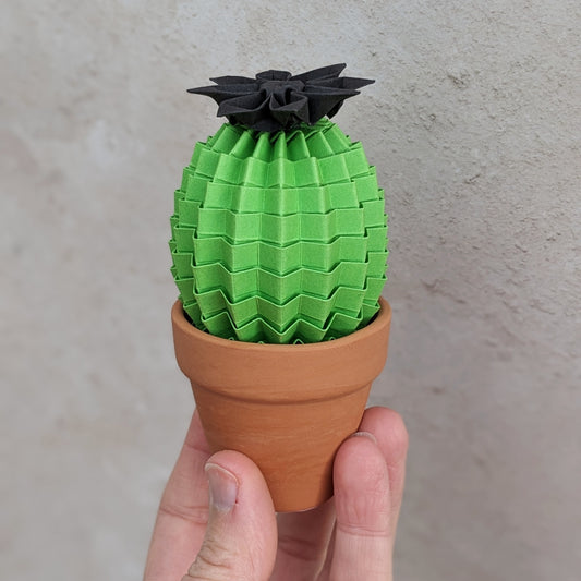 a hand holds a mini lime green origami paper cactus in a small terracotta pot. The cactus has a black paper flower on top