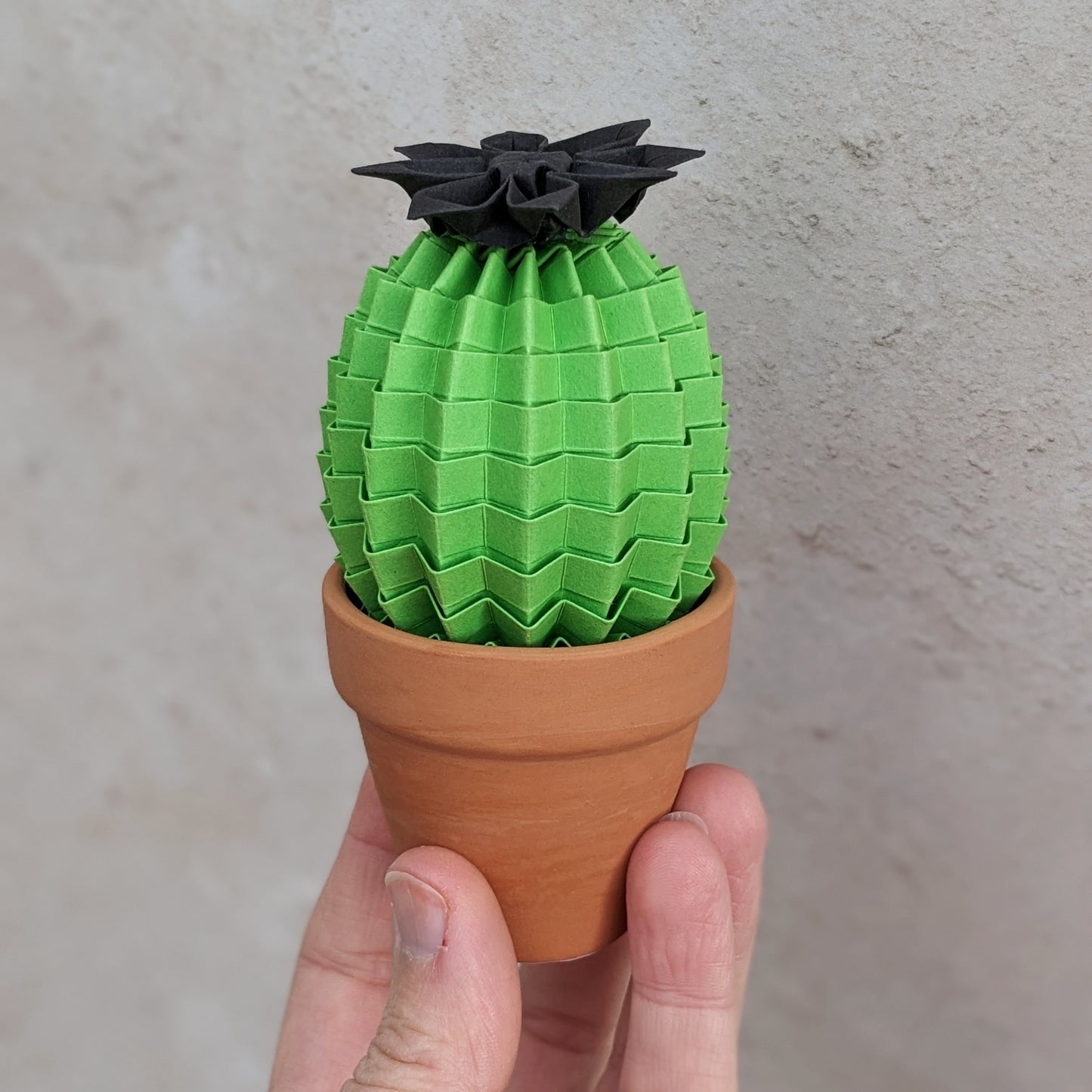 a hand holds a mini lime green origami paper cactus in a small terracotta pot. The cactus has a black paper flower on top